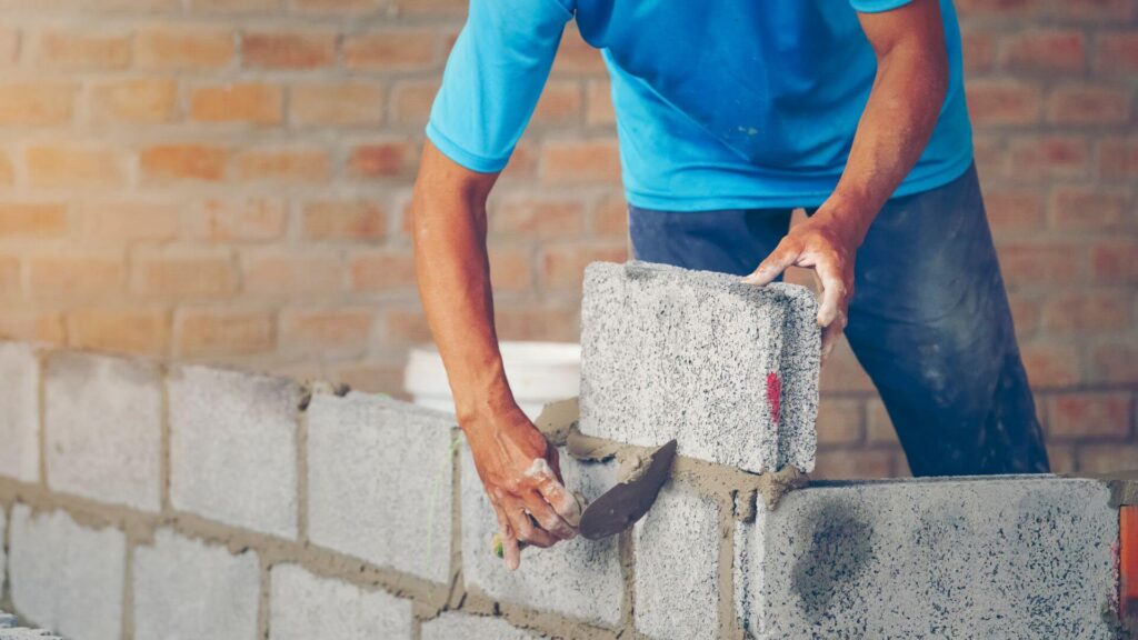 Pedreiro assentando blocos de concreto em parede de obra. Ilustra a fase de execução e a importância da qualidade dos materiais para evitar atrasos no cronograma.